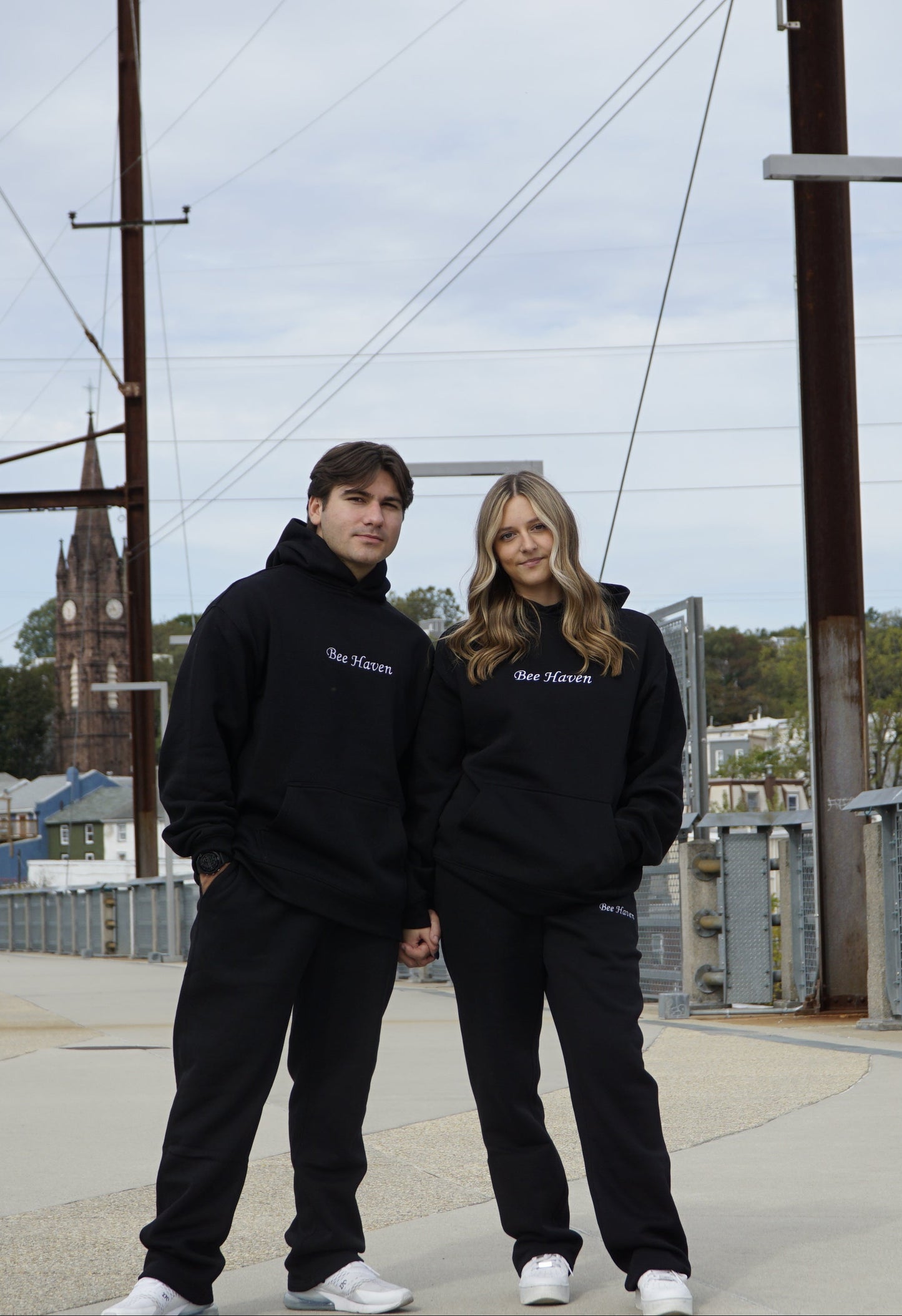 Two people wearing matching black hoodies on a rooftop with power lines and buildings in the background.