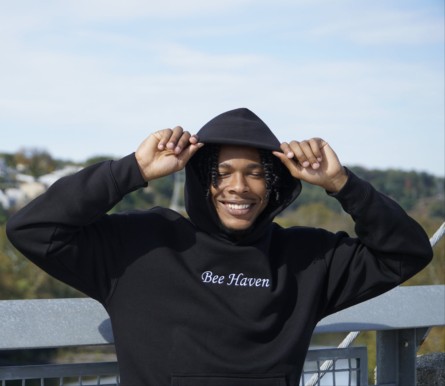 Person wearing a black hoodie with 'Bee Haven' text, standing outdoors with trees and buildings in the background.