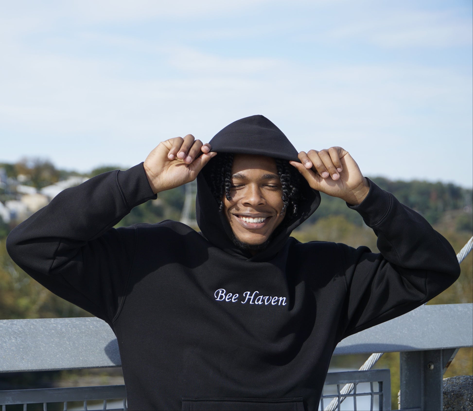 Person wearing a black hoodie with 'Bee Haven' text, standing outdoors with trees and buildings in the background.